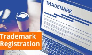 Hand holding magnifying glass over a trademark application form on a computer screen, surrounded by legal books and a framed certificate, highlighting the importance of trademark registration services for brand protection.