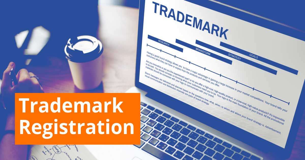 Hand holding magnifying glass over a trademark application form on a computer screen, surrounded by legal books and a framed certificate, highlighting the importance of trademark registration services for brand protection.